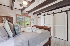 A bedroom with a wooden bed, a window with a view of greenery, a sliding door, and a ceiling fan. - Cozy NH Log Home | Ski, Hike & Unwind (Littleton)