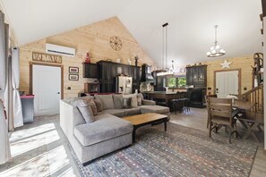 A spacious living room with a large sectional sofa, a wooden ceiling, and a kitchen area with dark cabinets and a chandelier. - Cozy NH Log Home | Ski, Hike & Unwind (Littleton)