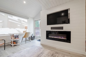 Un salon moderne avec une cheminée, un téléviseur fixé au mur et une fenêtre donnant sur des maisons enneigées.