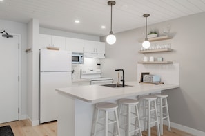 Une cuisine moderne avec des meubles blancs, un îlot central doté de tabourets, un four à micro-ondes, une cuisinière et un luminaire suspendu.