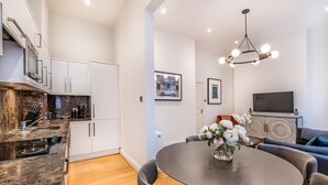 A modern kitchen with white cabinets, a dining table with chairs, and a living area with a sofa and TV. - Stylish Marylebone Apartment with Modern Soul (London)