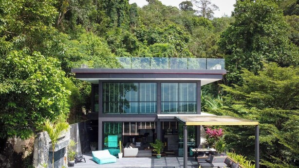 Una casa moderna de dos pisos con grandes ventanales, un patio con mobiliario exterior y abundante vegetación a su alrededor.