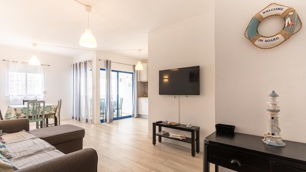 A modern living room with a TV, a dining area with chairs, a sofa, and a small table with a lamp. - 2 bedroom apartment Praia Da Rocha (Portimão)