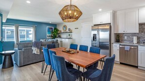 A modern kitchen with white cabinets, a stainless steel refrigerator, and a dining area with a wooden table and blue chairs. - 3BD/3BA | Ocean Views | 1 Block to the Beach (Oceanside)