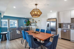 A modern kitchen with white cabinets, a stainless steel refrigerator, and a dining area with a wooden table and blue chairs. - 3BD/3BA | Ocean Views | 1 Block to the Beach (Oceanside)