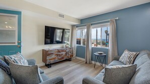 A modern living room with a TV, grey sofas, a wooden cabinet, and a view through a window. - 3BD/3BA | Ocean Views | 1 Block to the Beach (Oceanside)