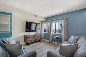 A modern living room with a TV, grey sofas, a wooden cabinet, and a view through a window. - 3BD/3BA | Ocean Views | 1 Block to the Beach (Oceanside)