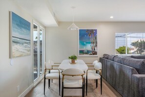 Een moderne woonkamer met een grijze bank, een ronde eettafel met stoelen, en een groot schilderij van een strandgezicht.