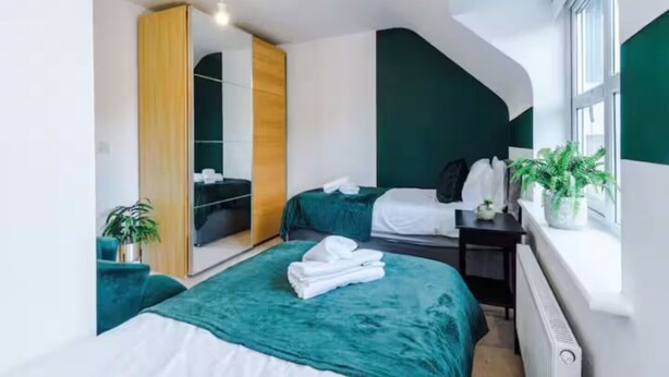 A modern bedroom with two beds, a large mirror, a wardrobe, and a window with a view.