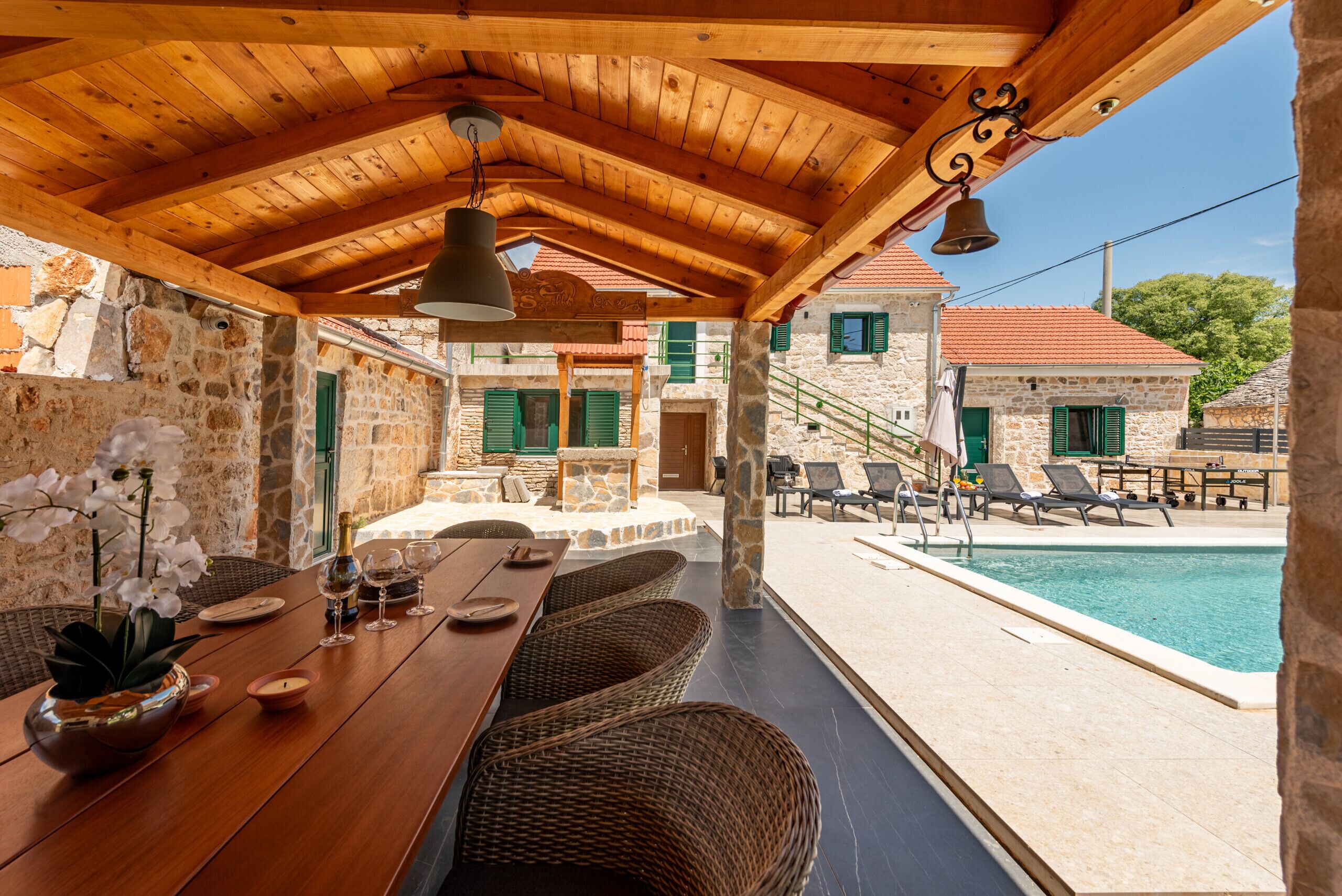 Un'area pranzo esterna in legno con pareti in pietra, una piscina e poltrona relax.