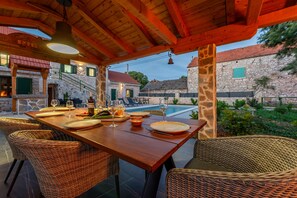 Un área de comedor al aire libre con sillas de mimbre y una mesa de madera dispuesta para una comida, con vistas a una piscina y edificios de piedra.