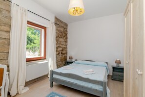 Un dormitorio con pared de piedra, una cama con cubrecama azul, una ventana con cortinas, una mesita de noche con lámpara y un armario de madera.