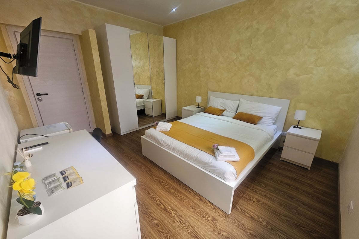 Una habitación de hotel con cama, mesitas de noche, un escritorio, un espejo y un televisor.
