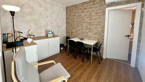 Een compacte kamer met een kitchenette, een eettafel met stoelen, een fauteuil, een lamp en een deur naar een andere kamer.