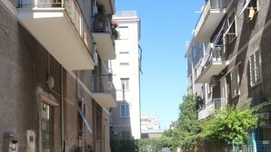 A narrow street flanked by multi-story residential buildings with balconies, potted plants, and a clear blue sky. - Empire home (Roma)