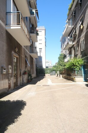 Una calle estrecha bordeada por edificios residenciales de varios pisos, con balcones y macetas con plantas, bajo un cielo azul despejado.