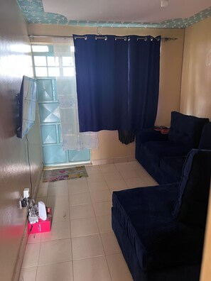 Ein Zimmer mit einem blauen Sofa, einem an der Wand befestigten Fernseher, einem Fenster mit Vorhängen und einem Fliesenboden.