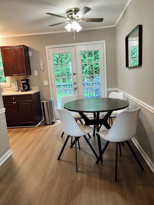 A dining area with a round table, four chairs, a ceiling fan, and a view through a glass door to a blue outdoor deck.