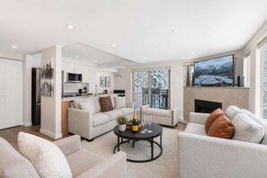 A modern living room with a sofa, coffee table, and a fireplace. There is a kitchen area with a microwave and a refrigerator. - Luxury 3-Bed Whistler Village | Private Hot Tub (Whistler)