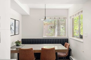 Een eetgedeelte met een glazen tafel, een beklede bank en hanglampen.