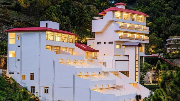 A modern multi-story hotel with a unique stepped design, large windows, and a red roof. - Misty Meadows Resort (Solan)