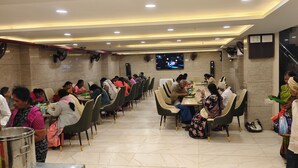 A dining area with multiple tables and chairs, a television screen mounted on the wall, and a staircase in the background. - Hotel Meritel Grande (Indore)