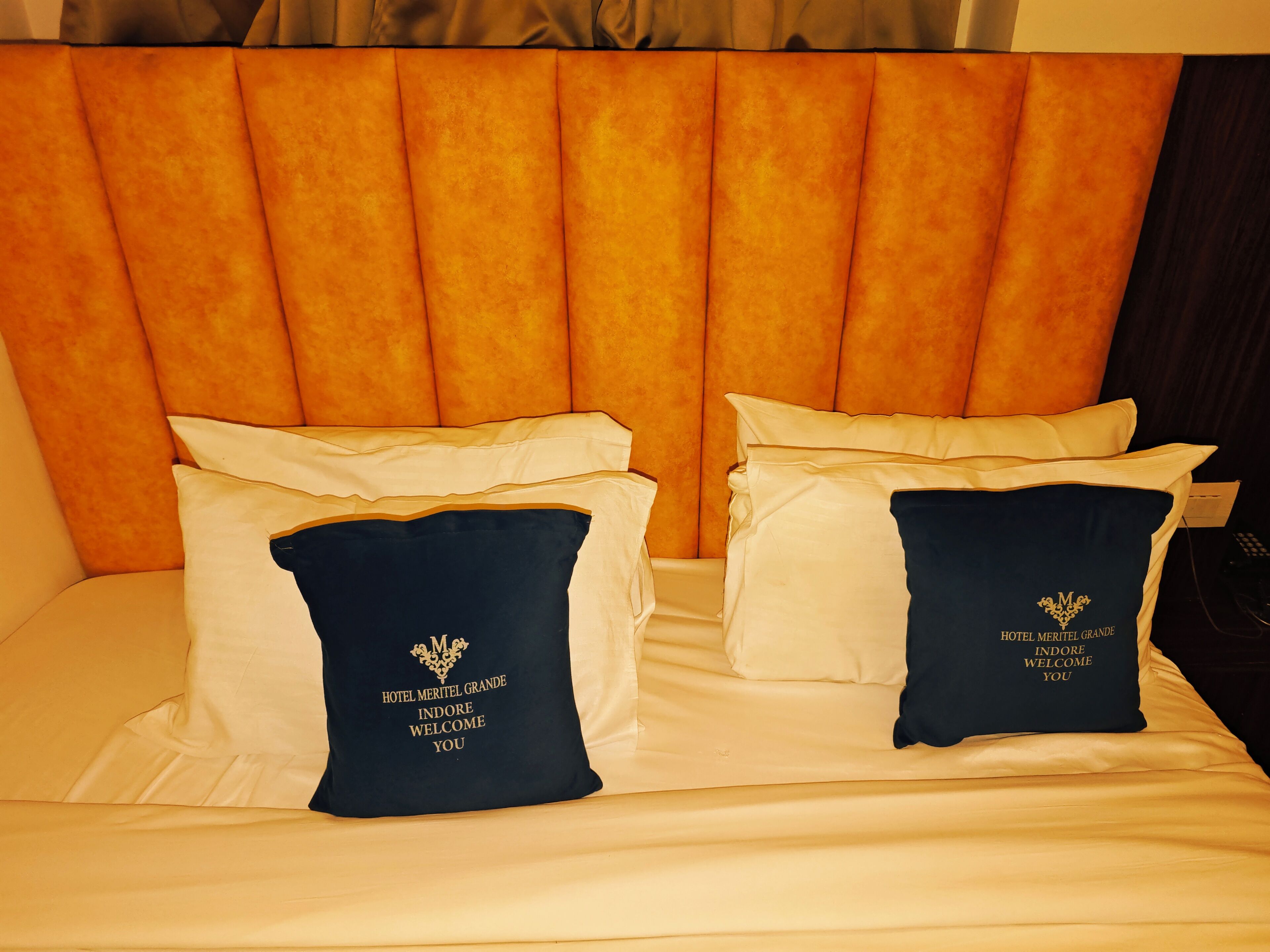 Ein Hotelbett mit gepolstertem Kopfteil, zwei Kissen mit dem Logo des Hotels und einer weißen Bettdecke.