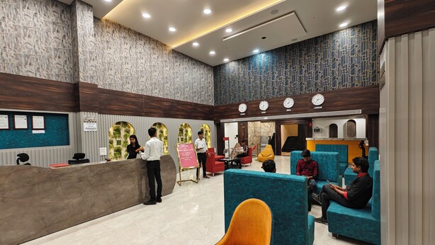 A hotel lobby with a reception desk, seating area, and a clock displaying time in multiple cities. - Hotel Meritel Grande (Indore)
