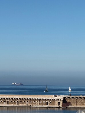 Blick auf die Küste mit einer Steinmauer, einem Segelboot und einem Frachter in der Ferne.