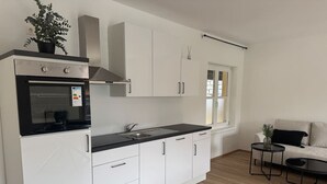 A modern kitchen with white cabinets, a black countertop, and a built-in oven. There is a window with blinds, a small round table, and a sofa in the adjacent living area. - Faraway Homes Studio Mautern #8 (Mautern in Steiermark)