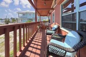 Eine überdachte Veranda mit Korbstühlen, einem Holzbalkon und Blick auf ein blaues Haus im Hintergrund.