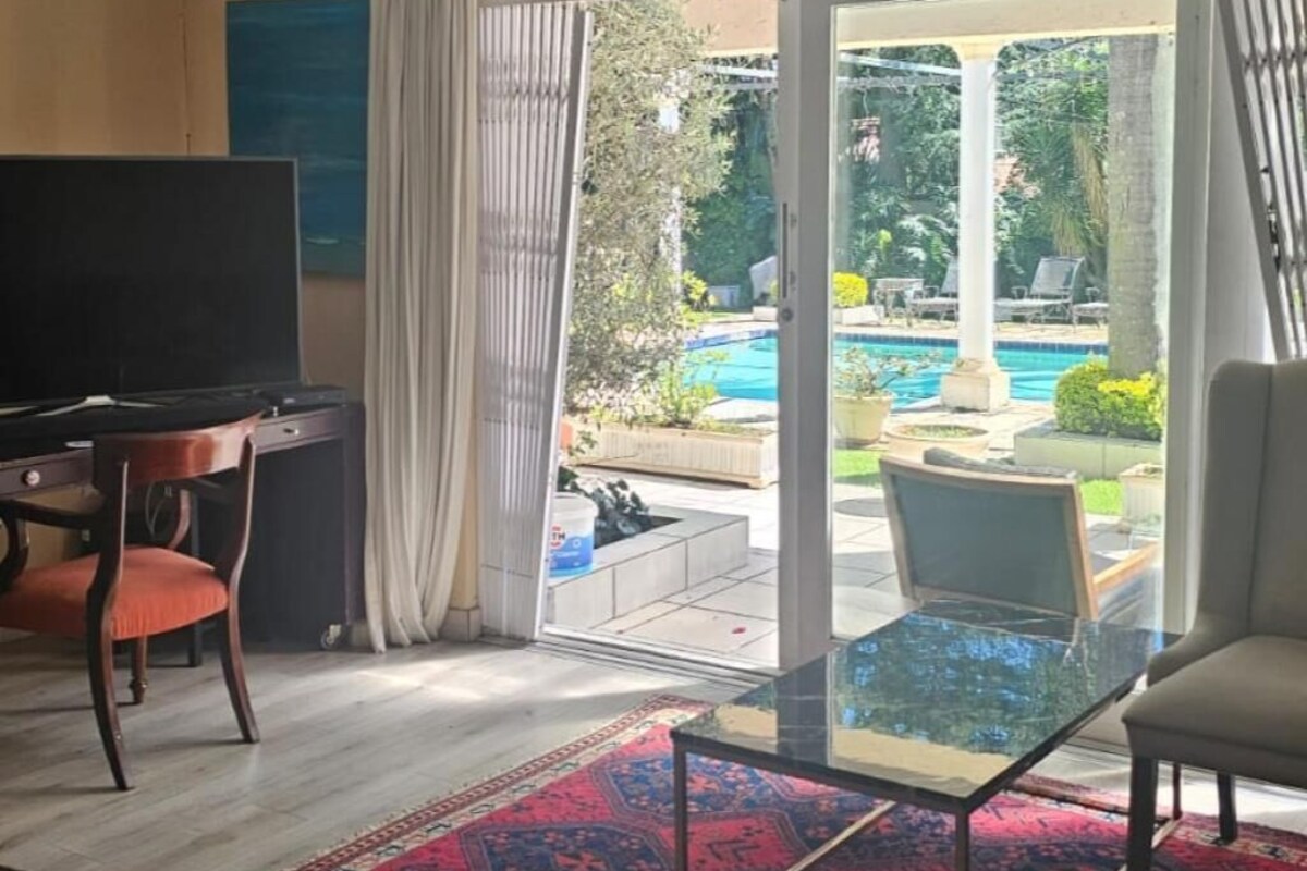A living room with a TV, a glass table, a sofa, and a view of a pool and garden.