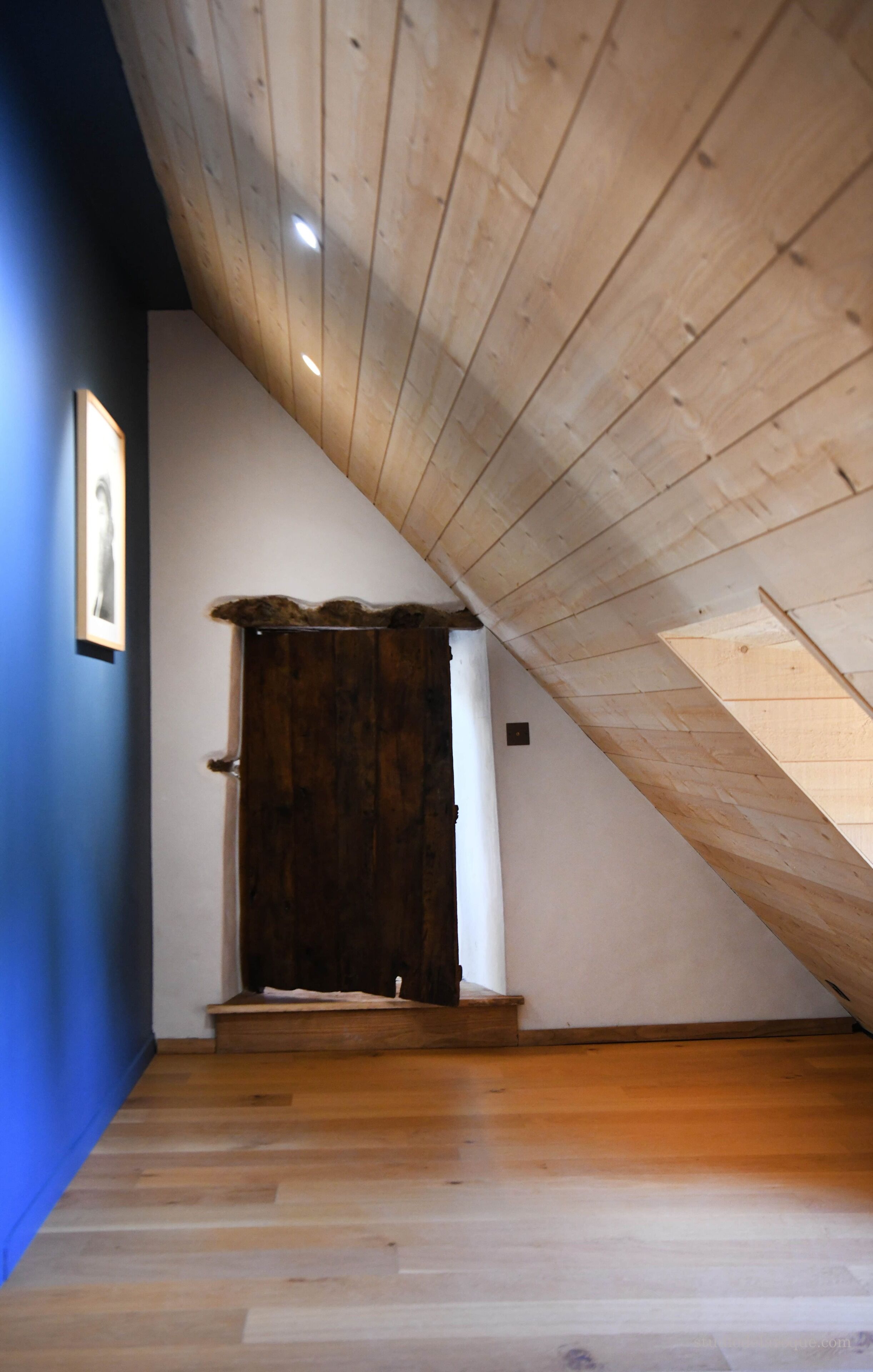 Una habitación con techo inclinado, una puerta de madera y un cuadro en la pared.