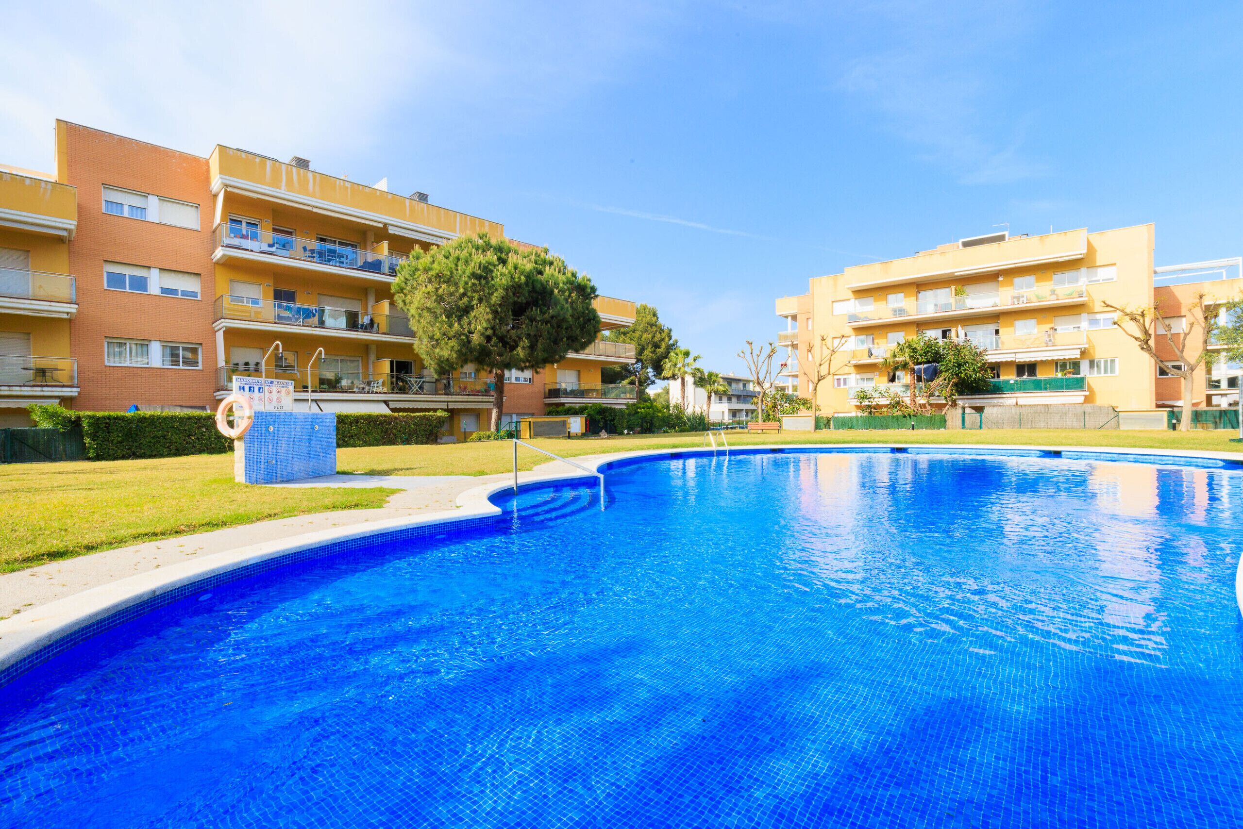 A swimming pool with a tiled edge, surrounded by a grassy area and multi-story buildings.