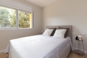Een slaapkamer met een bed, kussens, een nachtkastje, een lamp en een raam met uitzicht op bomen.