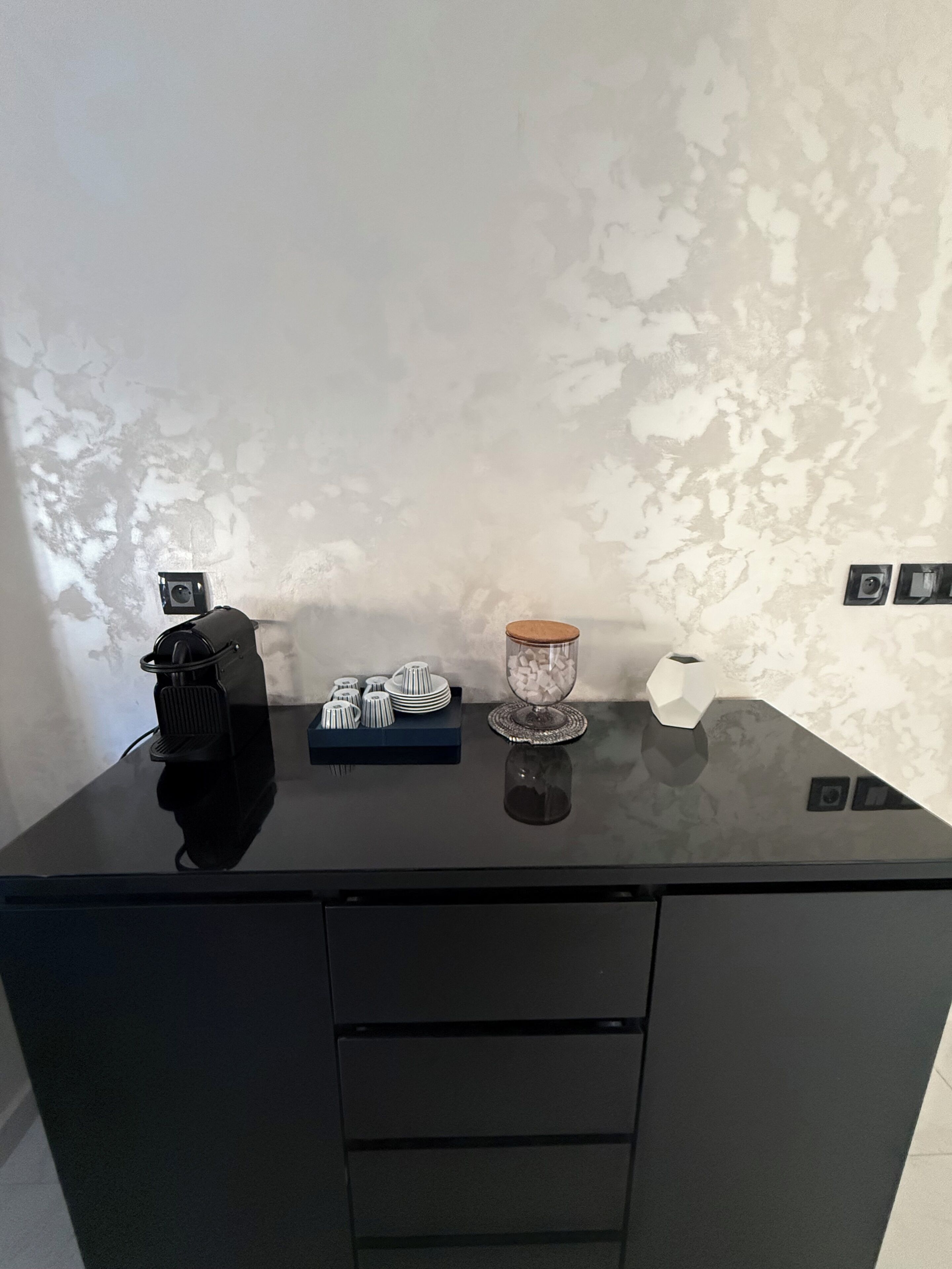 Un escritorio negro con un altavoz, un recipiente de vidrio con cubos y un objeto blanco sobre una pared con textura.