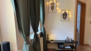 Une pièce avec une fenêtre, des rideaux, une petite table ornée d’objets décoratifs et un mur où sont accrochées des œuvres d’art encadrées.