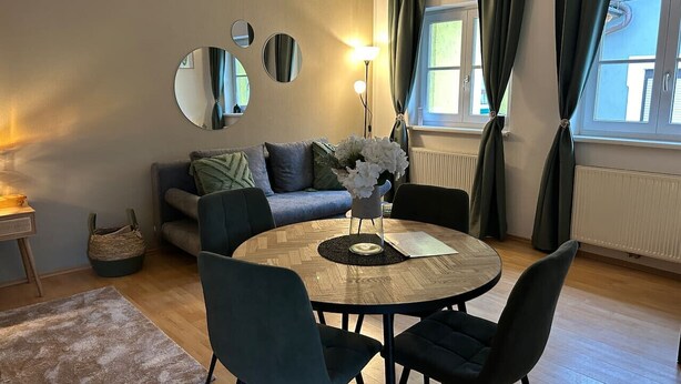 Un salon chaleureux avec une table ronde en bois, des fauteuils foncés, un canapé avec des coussins, une table d’appoint et un miroir au mur.