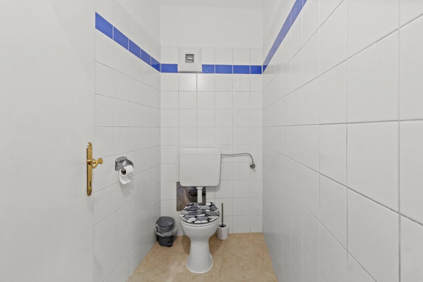 A small, white-tiled bathroom with a toilet, a trash bin, and a wall-mounted air vent.