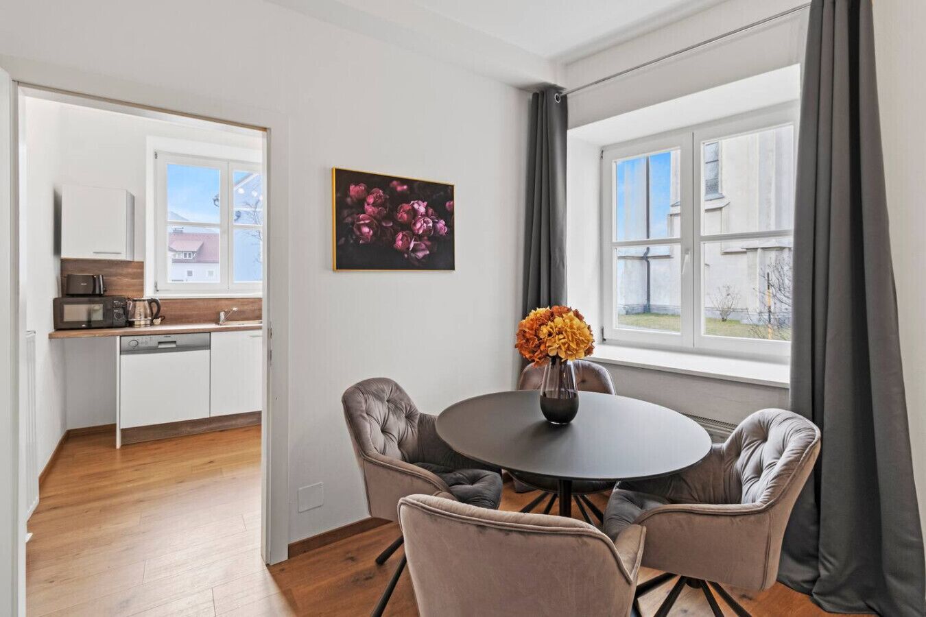 A modern kitchen with white cabinets, a microwave, and a coffee maker. There is a dining area with a round table and four chairs, a vase with flowers, and a window with curtains.