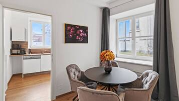 A modern kitchen with white cabinets, a microwave, and a coffee maker. There is a dining area with a round table and four chairs, a vase with flowers, and a window with curtains.
