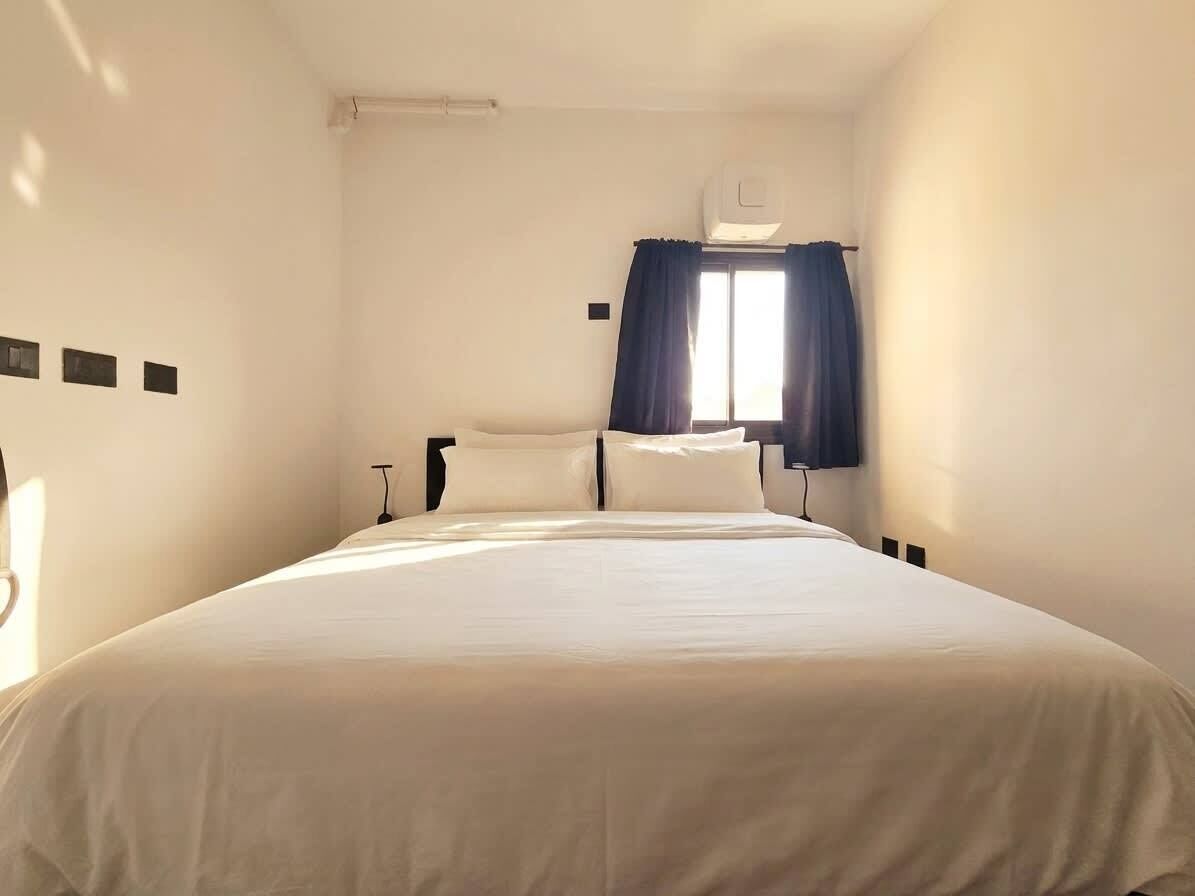Habitació d'hotel amb llit, finestra amb cortines, aire condicionat fixat a la paret i lluminària penjant de la paret.