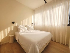 A bedroom with a large bed, a bedside table, and a window with sheer curtains.