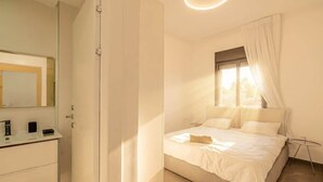Ein Schlafzimmer mit Bett, Nachttisch, einem Fenster mit Vorhängen, einem Spiegel und einem Badezimmer mit Waschbecken und Handtuchhalter.