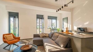 Ein modernes Wohnzimmer mit einer Couch, einem Sessel, einem Couchtisch und einer Küchenzeile mit Arbeitsfläche und Geräten.