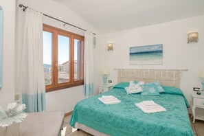 Une chambre à coucher avec un lit, des rideaux, une table de chevet, une lampe, un tableau et une vue sur les bâtiments à l’extérieur.