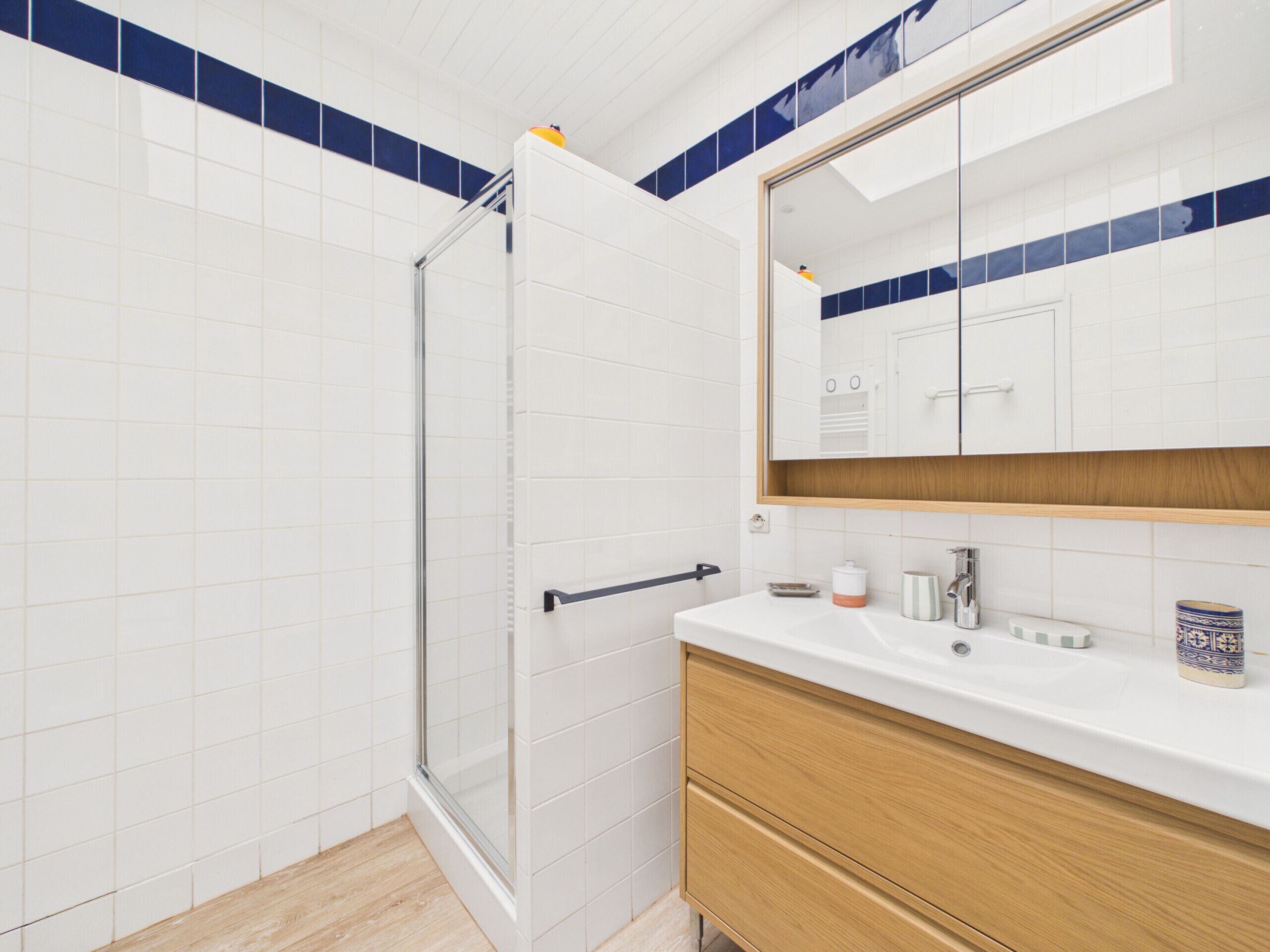 Baño con azulejos blancos, cabina de ducha de vidrio, tocador de madera y espejo sobre el lavabo.