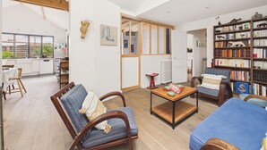 Amplia sala de estar con sofá, sillón y mesa de centro, una estantería y una cocina visible a través de una puerta abierta.