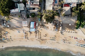Eine Vogelperspektive eines Strandes mit Reihen von Sonnenschirmen, Liegestühlen und einigen Gebäuden im Hintergrund.