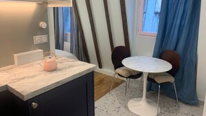 Una cocina compacta con encimera de mármol, una mesita, dos sillas, una cesta colgante y un ventanal con cortinas.